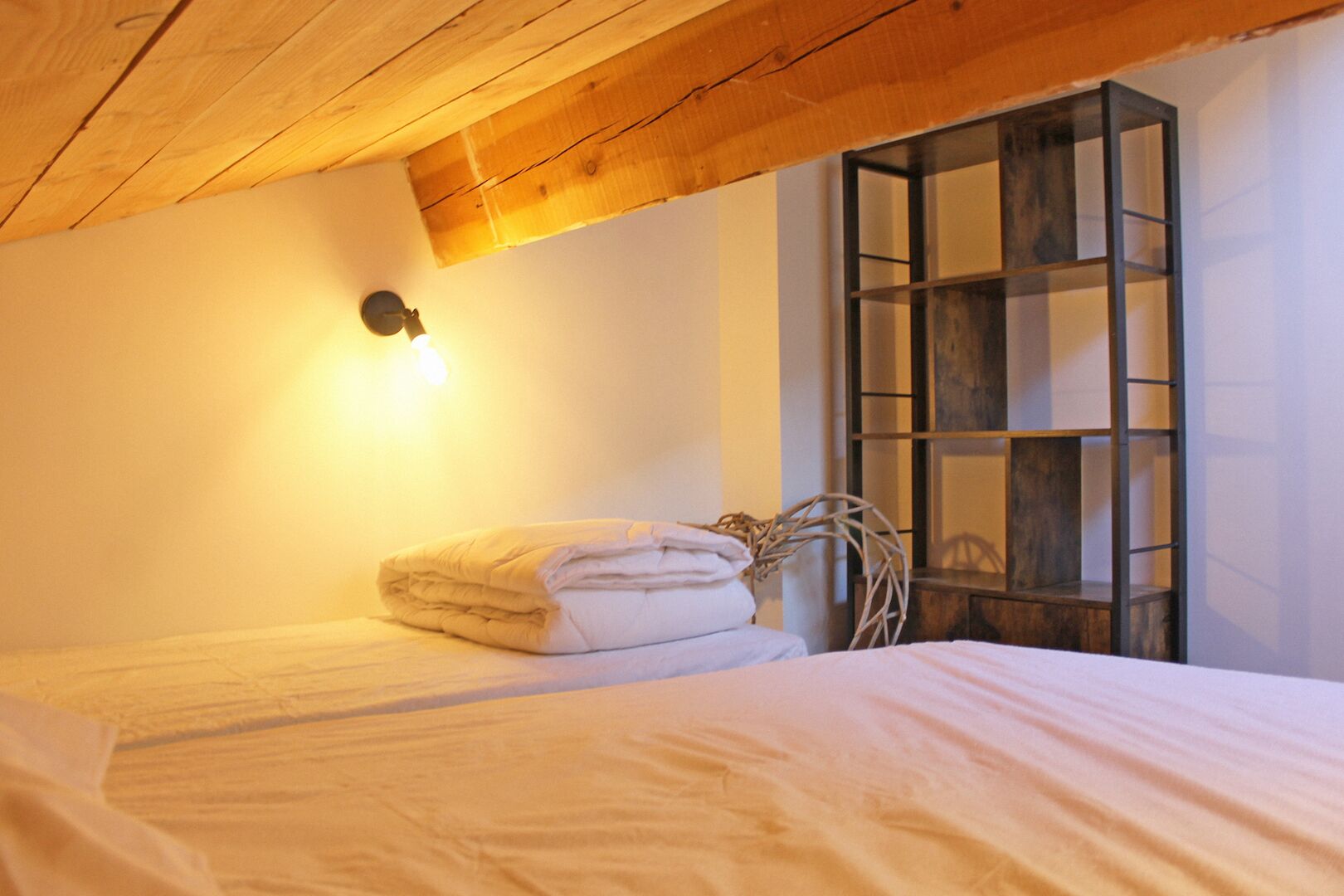Un dormitorio acogedor con techo de madera, una cama con ropa de cama blanca impecable, una lámpara de pared y una estantería de escalera de madera.