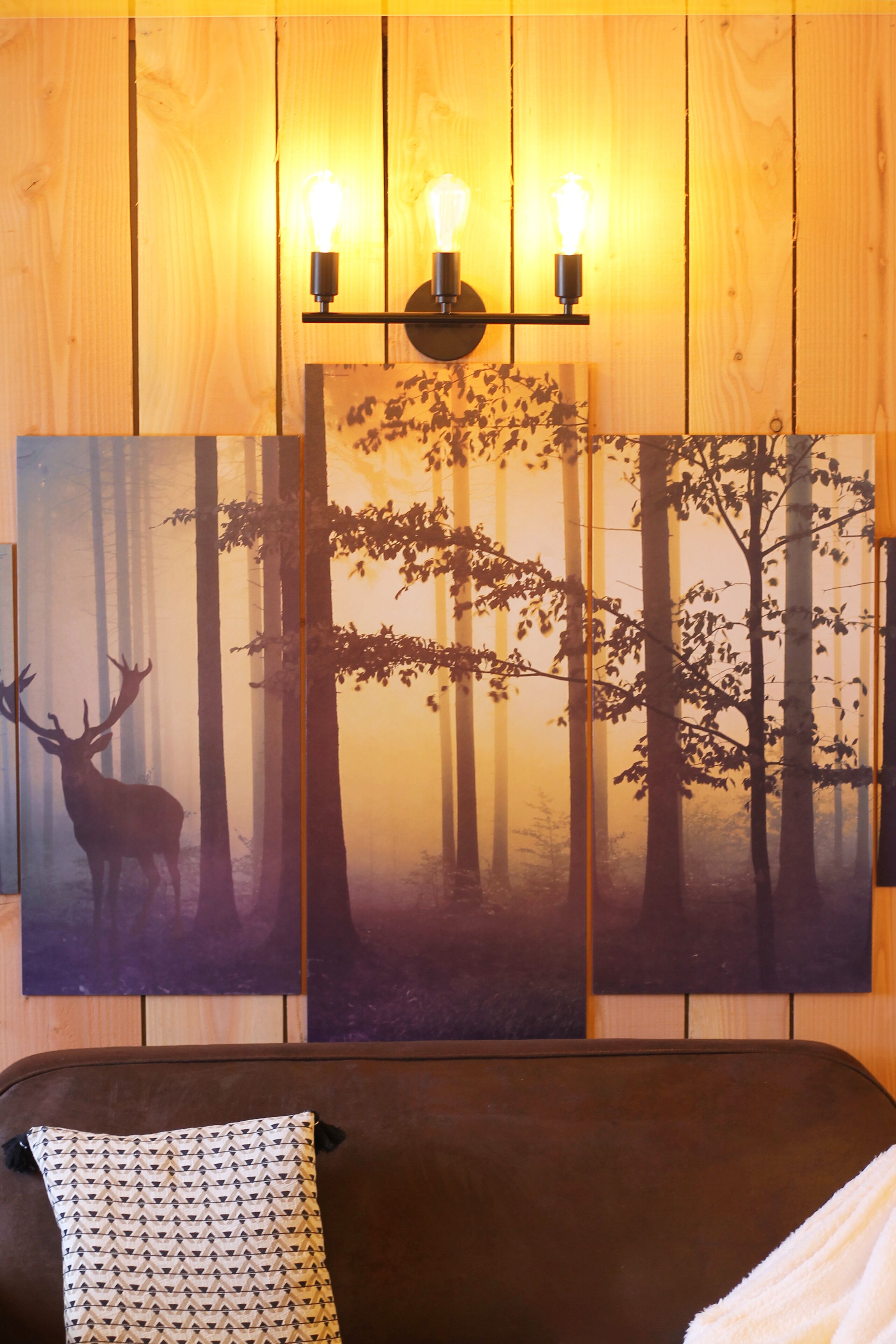A room with a large wall-mounted painting of a forest scene, a brown sofa, and a patterned pillow.