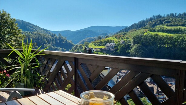 A wooden deck with a table, a glass with a drink, and a view of green hills and buildings.