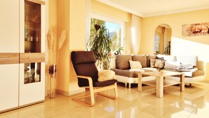 A modern living room with a sofa, armchairs, and a coffee table. There is a large window with plants and a view of the outdoors.
