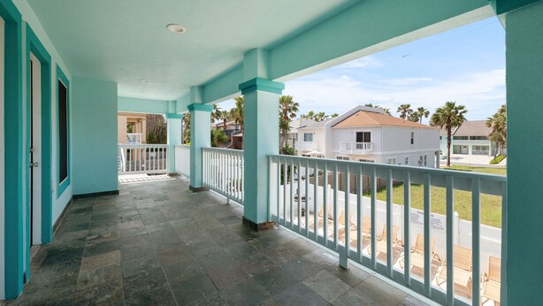 A covered balcony with teal columns and white railings, overlooking a residential area with palm trees and houses. - Lantana Retreat 3 - Spacious 3BR w/ Pool (South Padre Island)