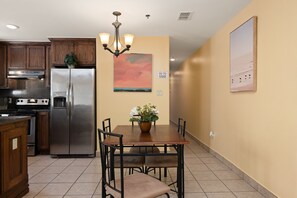 A kitchen with wooden cabinets, a stainless steel refrigerator, a dining table with chairs, a chandelier, and a painting of a beach scene.