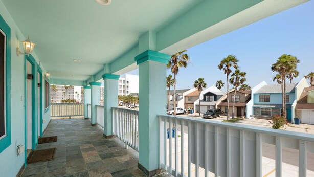 A covered balcony with teal columns and a stone floor, overlooking a street with palm trees and buildings.