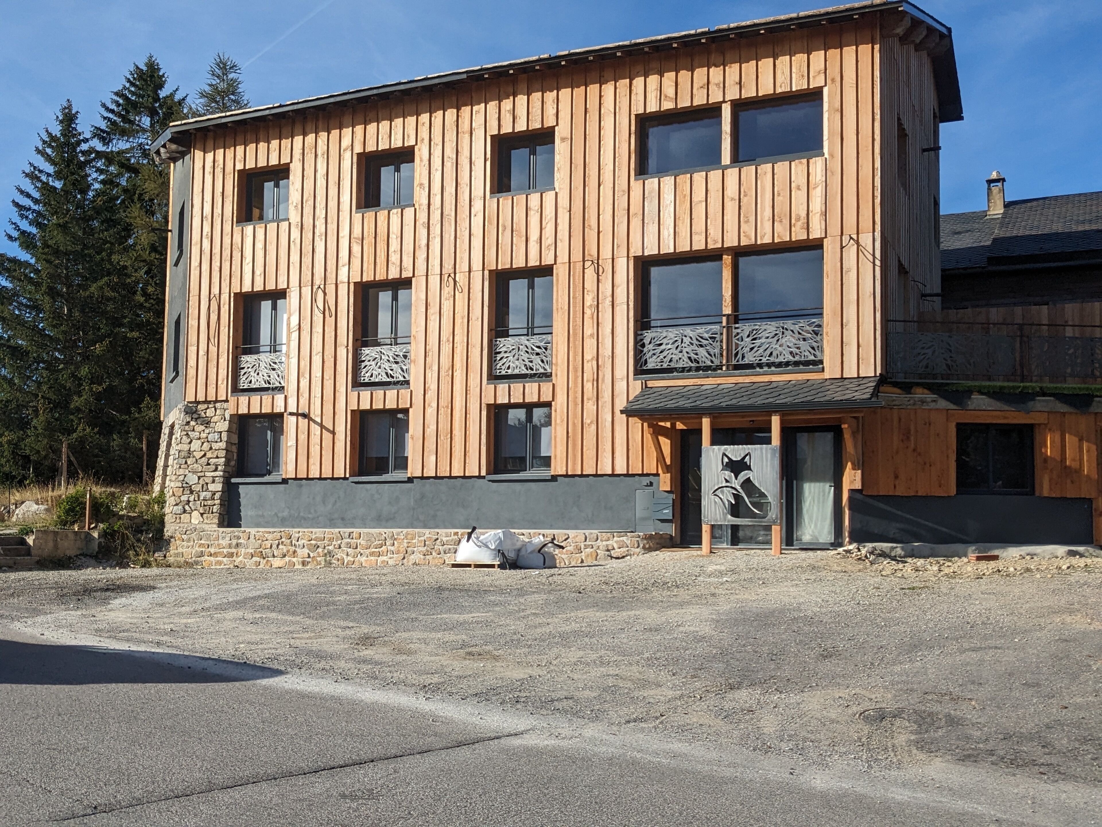 A modern wooden building with multiple windows and balconies, stone foundation, and a gravel driveway.