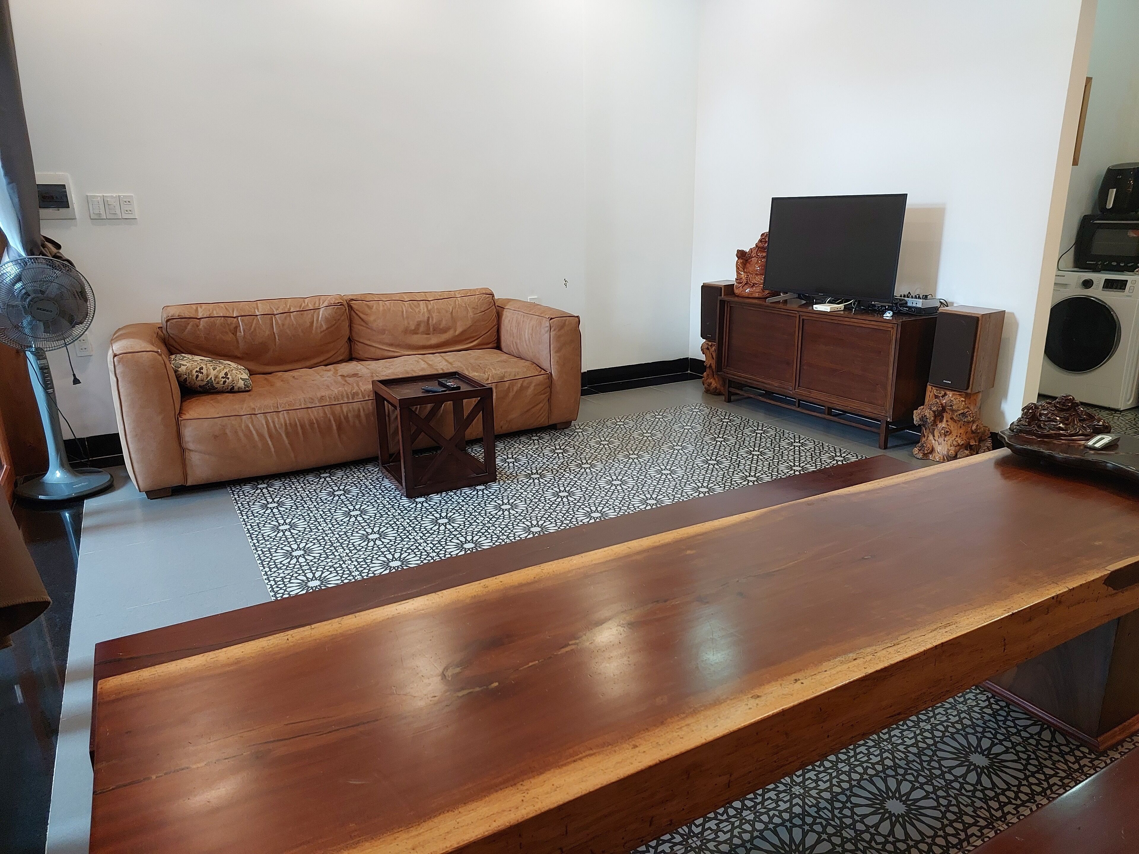 A living room with a brown leather sofa, a small wooden coffee table, a TV on a wooden cabinet, and a large wooden dining table.
