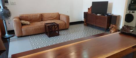 A living room with a brown leather sofa, a small wooden coffee table, a TV on a wooden cabinet, and a large wooden dining table.