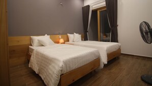 Un dormitorio con dos camas, cabeceros de madera, una mesita de noche con lámpara, una ventana con cortinas y un ventilador de pared.