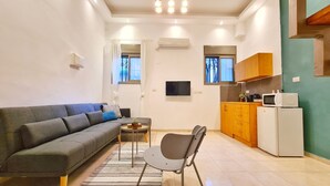 A compact living space with a sofa, a small dining table, a chair, a kitchen area with cabinets and appliances, and a flat-screen TV mounted on the wall. - Bugrashov Beach Studios (Tel Aviv)