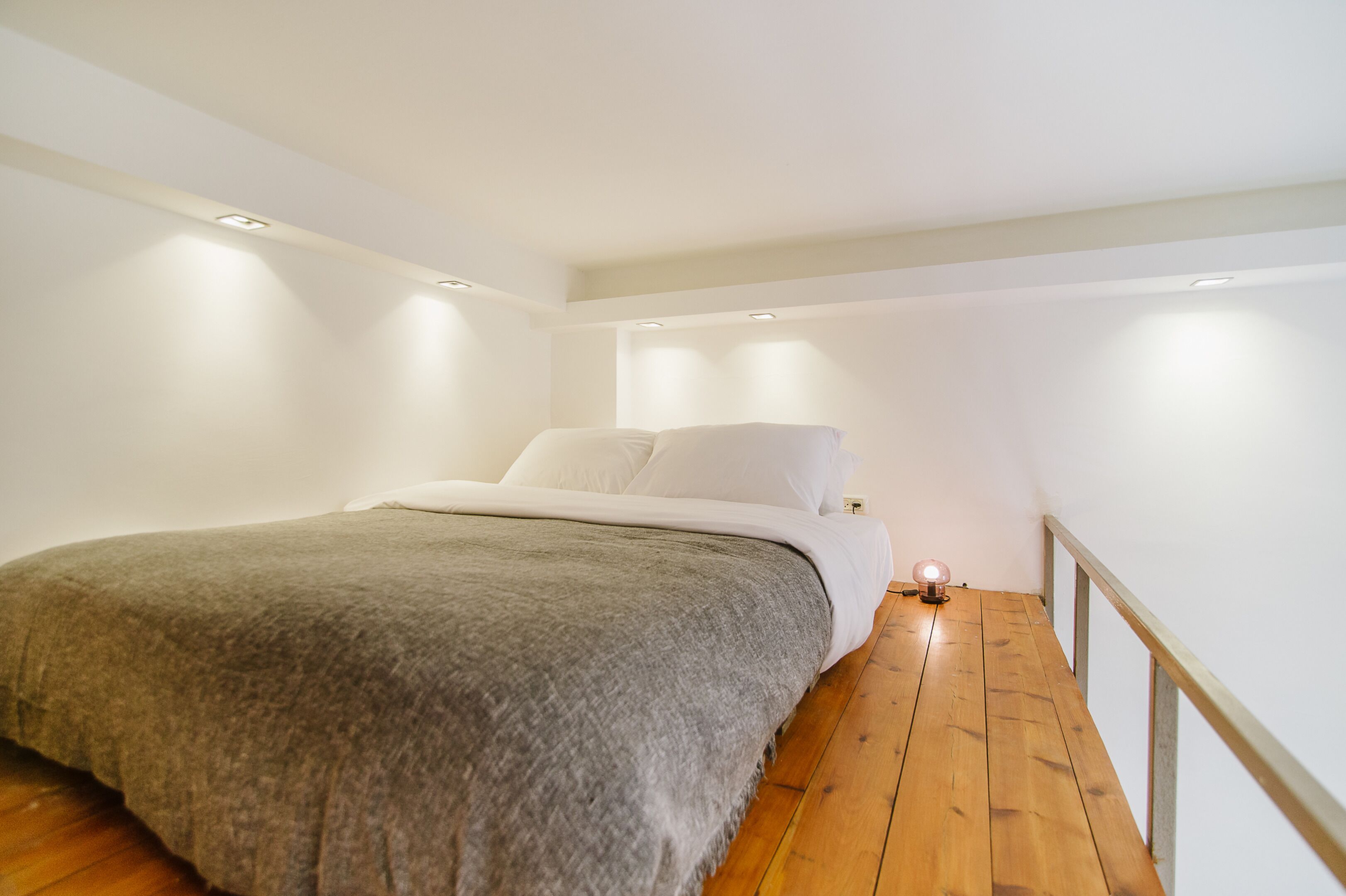 A bedroom with a bed, a bedside table with a lamp, wooden flooring, and white walls.
