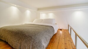 A bedroom with a bed, a bedside table with a lamp, wooden flooring, and white walls. - Bugrashov Beach Studios (Tel Aviv)