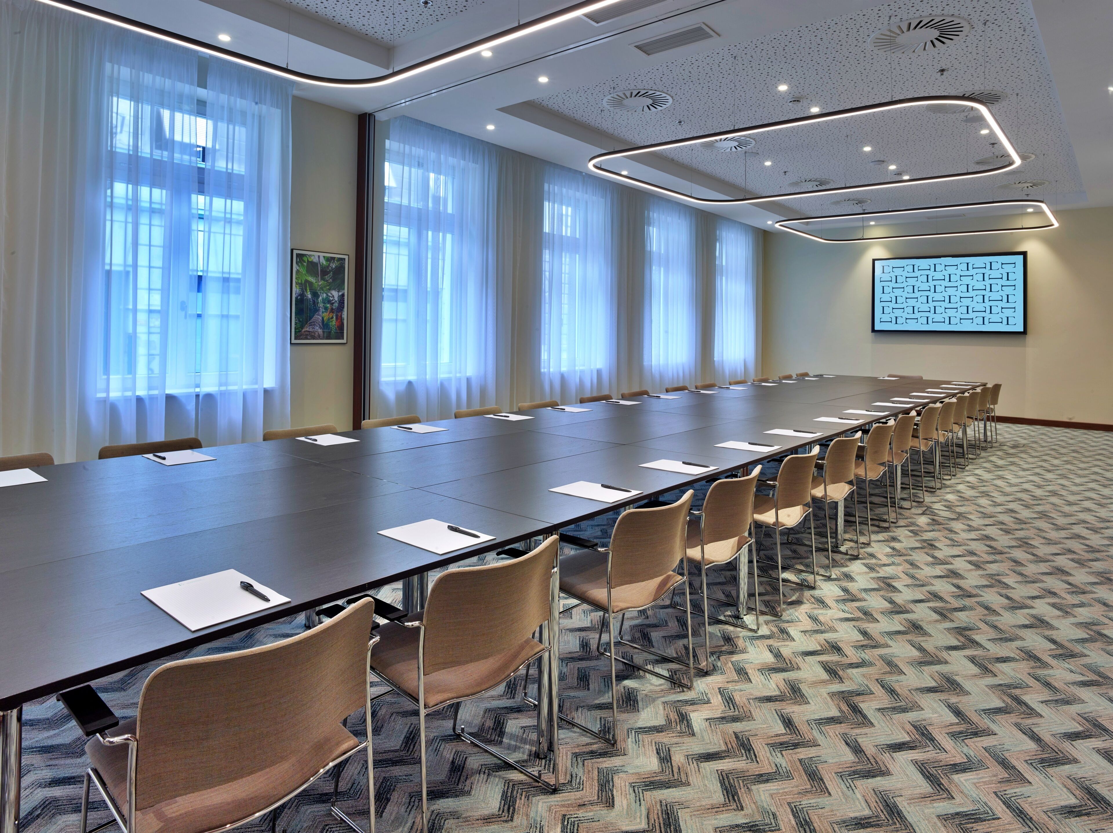 Una sala de conferencias con una larga mesa, sillas, una pantalla de proyector y ventanas grandes con cortinas.