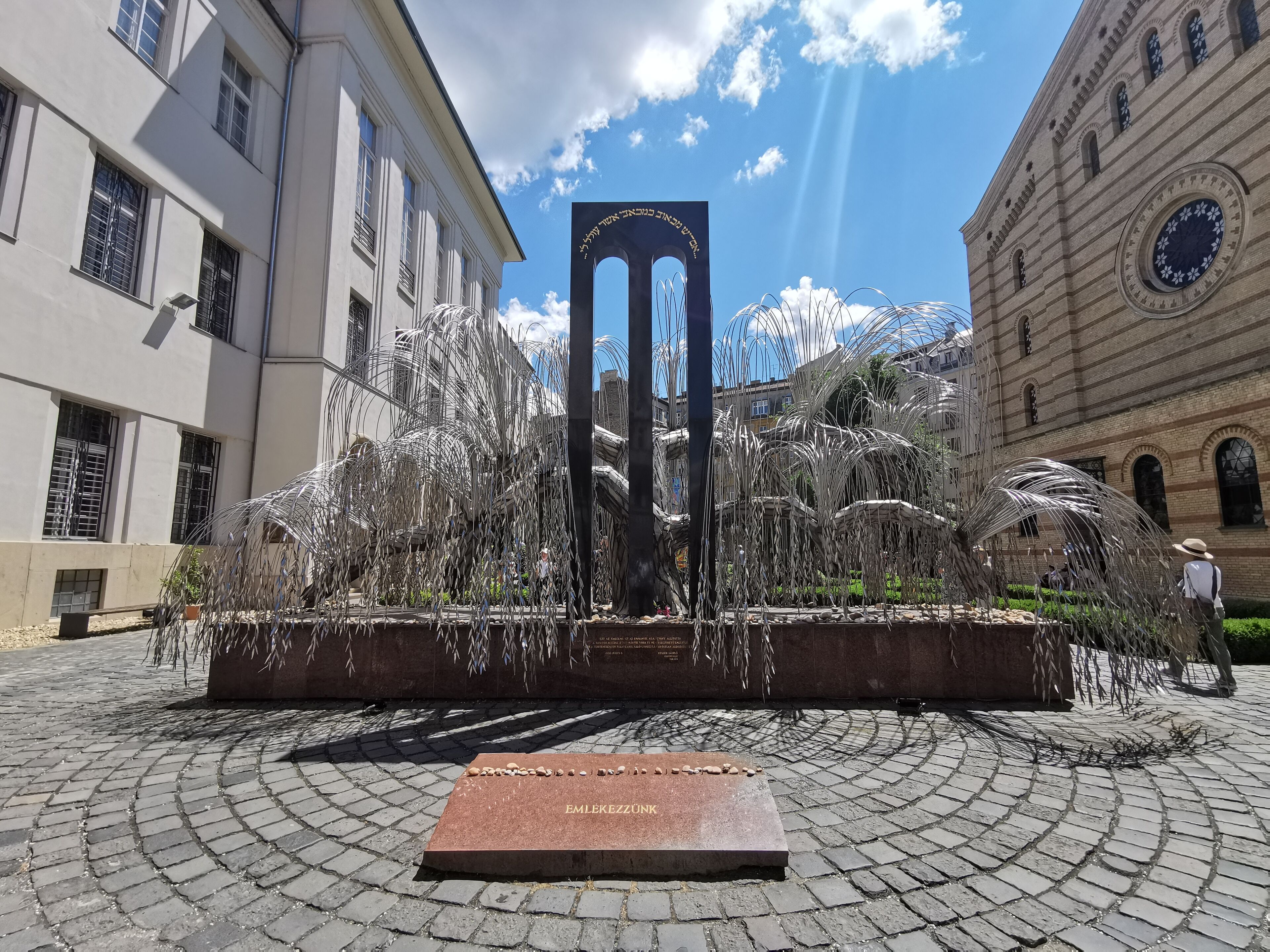 A public square with a central sculpture, cobblestone pavement, and surrounding buildings.