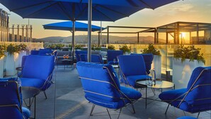 Terraza en la azotea con sillas y sombrillas azules, que ofrece vistas del horizonte urbano al atardecer.