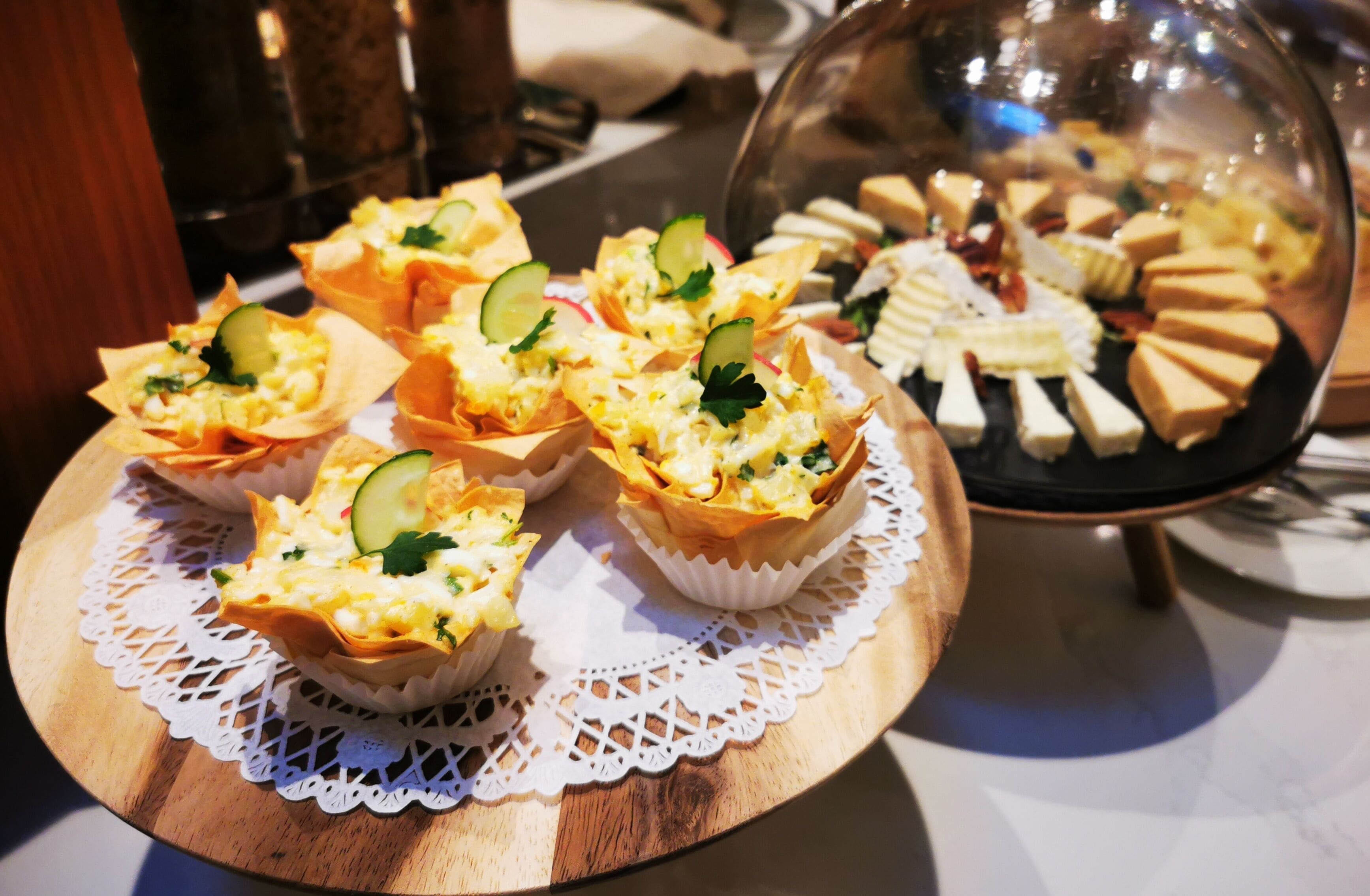 A variety of appetizers on a wooden board with a lace doily, including some with cucumber and cheese.