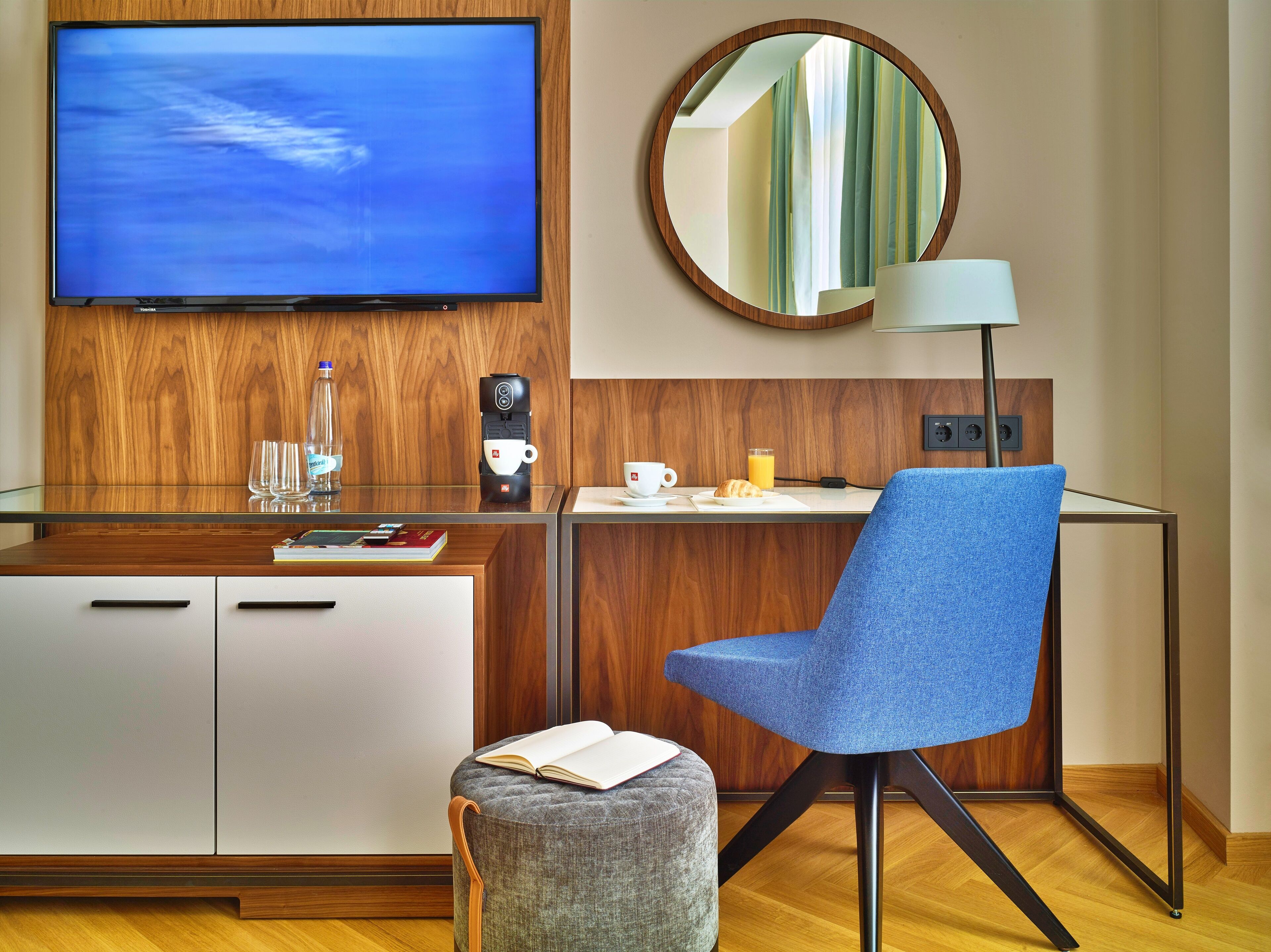A modern hotel room with a wooden desk, a blue chair, a round mirror, a flat-screen TV, and a coffee machine.