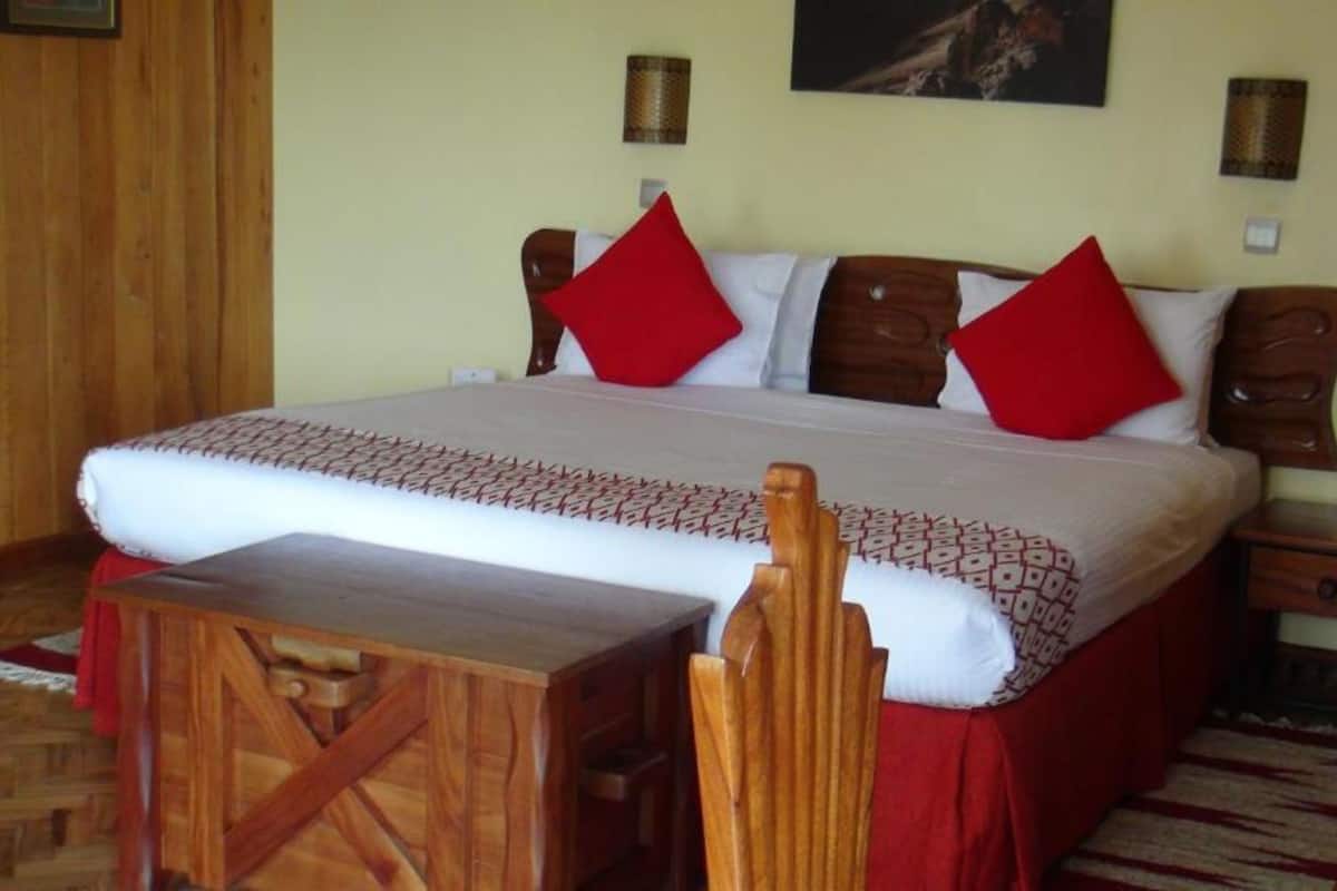 A neatly made bed with a patterned bedspread and red pillows, a wooden nightstand with a drawer, and a small table with a chair in a room with wooden flooring and a red rug.