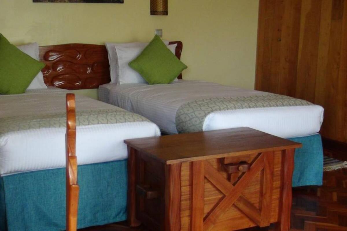 A hotel room with two beds, each with a wooden headboard, green pillows, and blue bedspreads. There is a wooden nightstand in front of the beds.