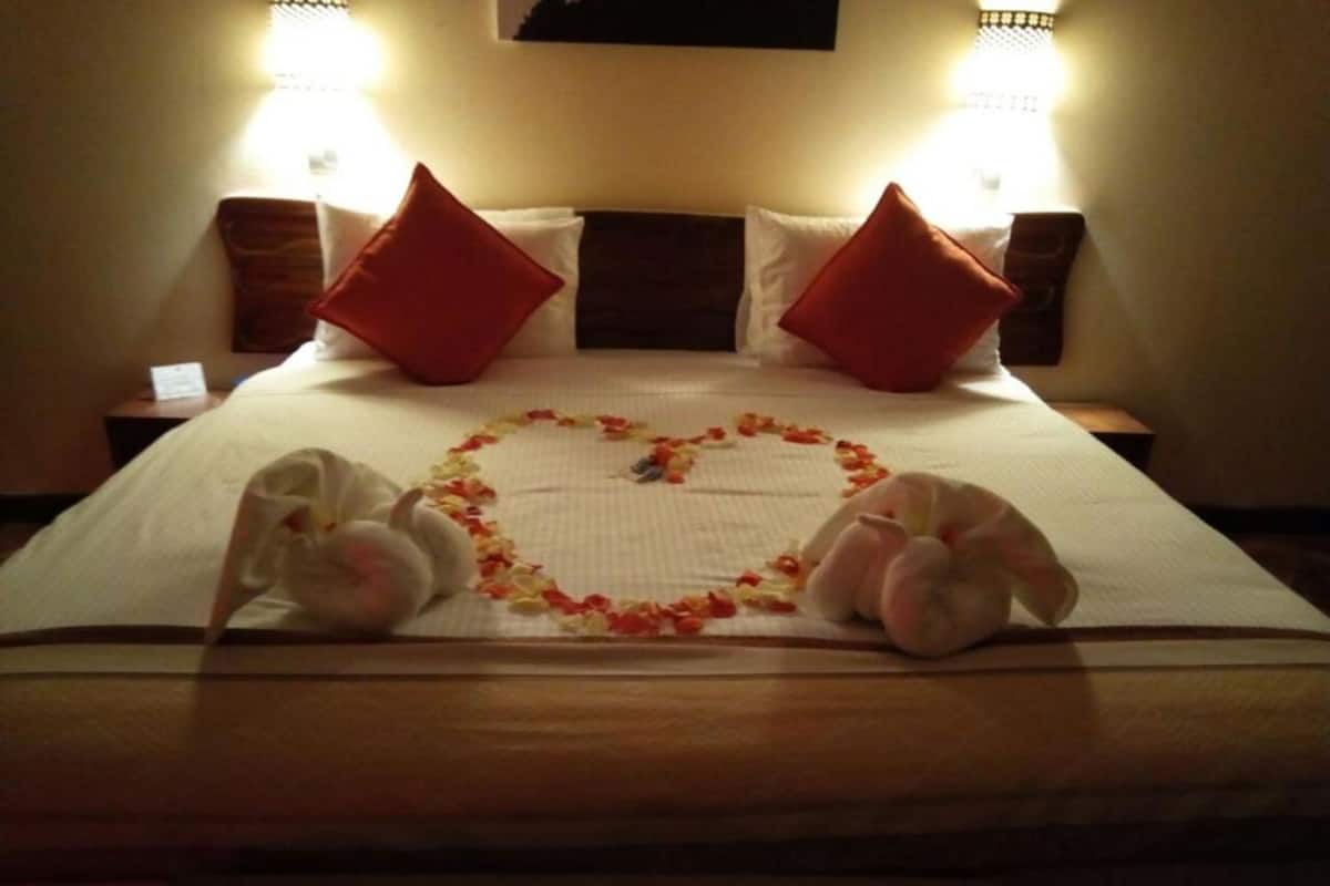 A hotel room with a bed, two red pillows, a heart-shaped arrangement of flowers, and two white towels.