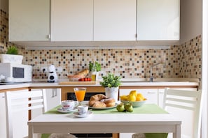 A kitchen with white cabinets, a tiled backsplash, and a breakfast table set with croissants, a bowl of fruit, and a coffee maker.