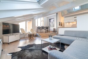 A modern living room with a large TV, grey sofa, and a dining area with a table and chairs. - Haus La Rocca by Zermatt Premium Apartments (Zermatt)