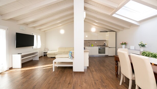 Un vaste espace de vie doté d’un haut plafond, d’un plancher en bois, d’un téléviseur à écran plat, d’un canapé, d’une table à manger avec des chaises, et d’une cuisine à l’arrière-plan.