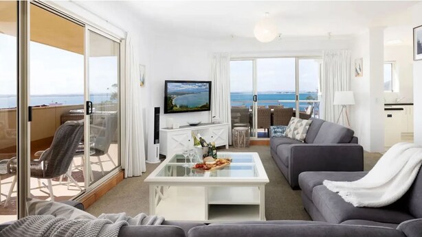 Ein modernes Wohnzimmer mit Sofa, Couchtisch und Fernseher. Ein Balkon bietet Blick auf das Meer.