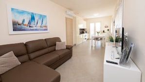 A modern living room with a brown sofa, a painting of sailboats on the wall, a kitchen area in the background, and a dining table with chairs. - Appartamento Lilli (Manerba del Garda)
