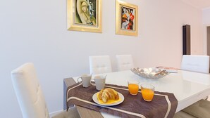 Un comedor con una mesa blanca, dos obras de arte enmarcadas, un croissant, un vaso de jugo de naranja y un bol de fruta.