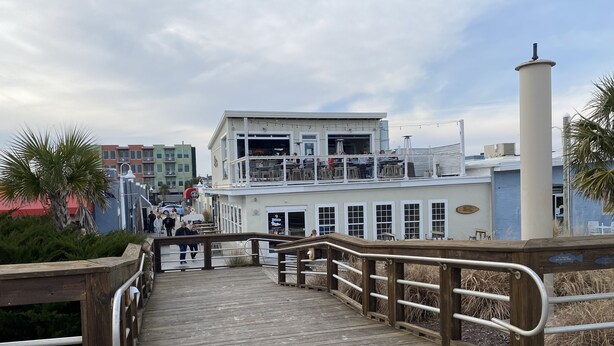 A wooden boardwalk leading to a waterfront building with outdoor seating and a view of the ocean.