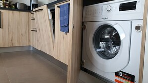 A compact kitchen with a built-in Whirlpool washing machine, wooden cabinets, and a shelf with kitchen items. - Le petit marquisat à deux pas du lac (Annecy)