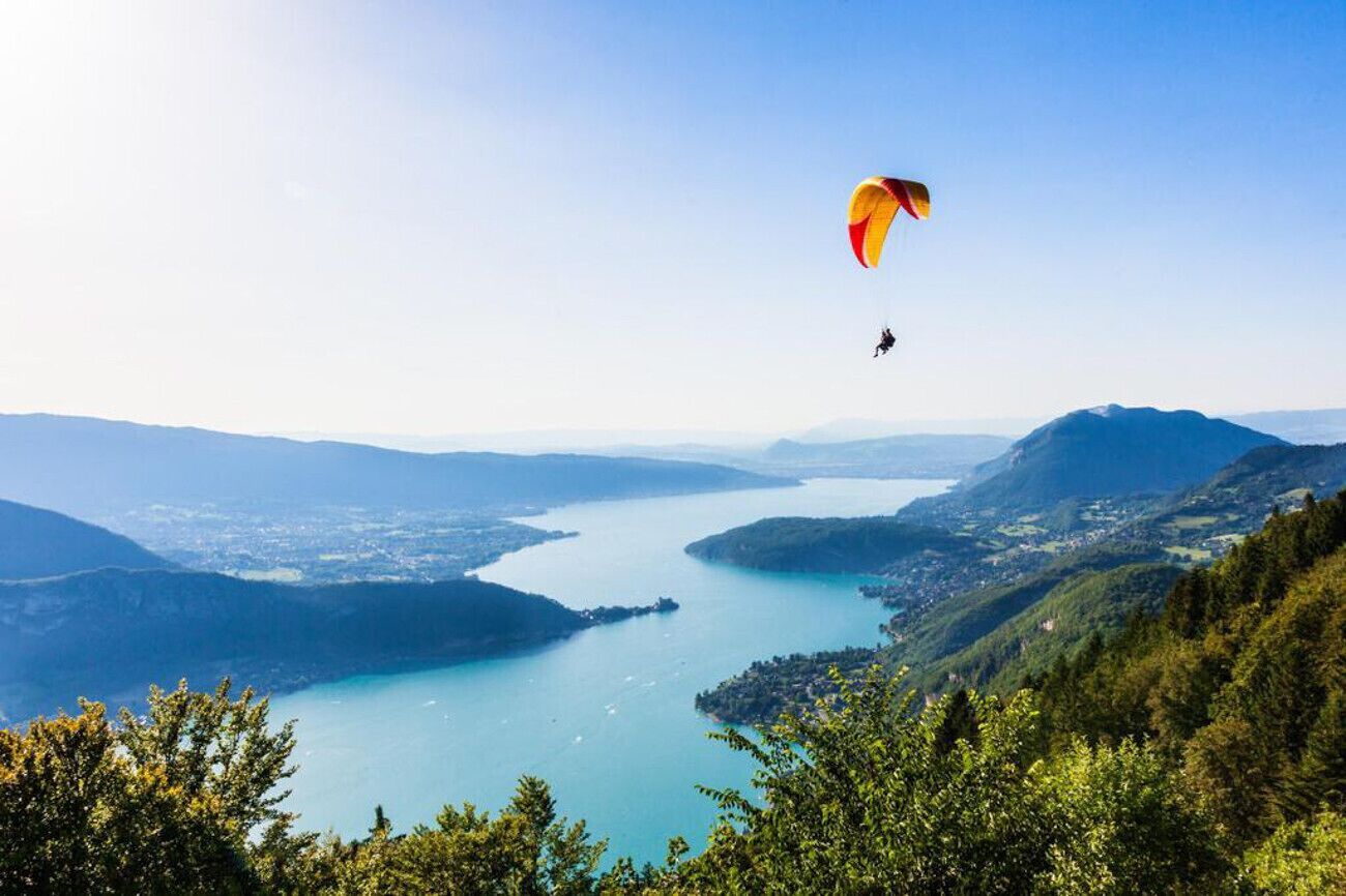 Una persona practica parapente sobre un lago, rodeado de colinas y un cielo despejado.