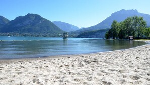 A beach with a clear view of mountains in the background, a calm lake, and a small structure on the water.