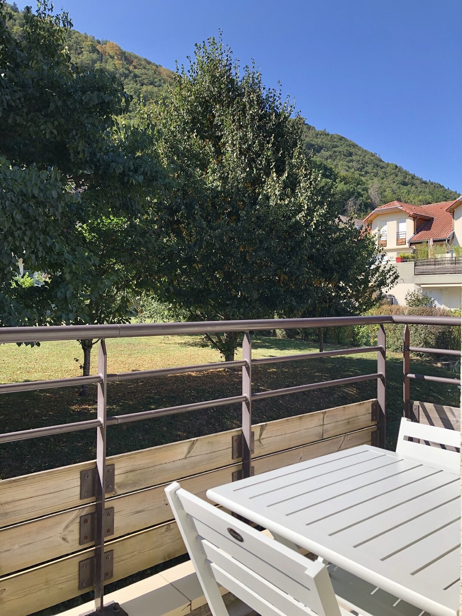 A balcony with a wooden table and chairs, overlooking a grassy area and a hill with trees.
