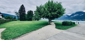 A well-maintained grassy area with a paved path, a large tree, and a view of a lake with mountains in the background.