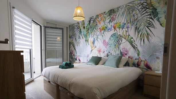 Camera da letto con un letto grande, una porta scorrevole in vetro, un murale tropicale e una lampada a sospensione.