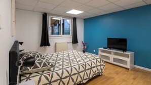 Une chambre à coucher avec un lit, un téléviseur posé sur un meuble blanc, une fenêtre avec des rideaux et un radiateur.