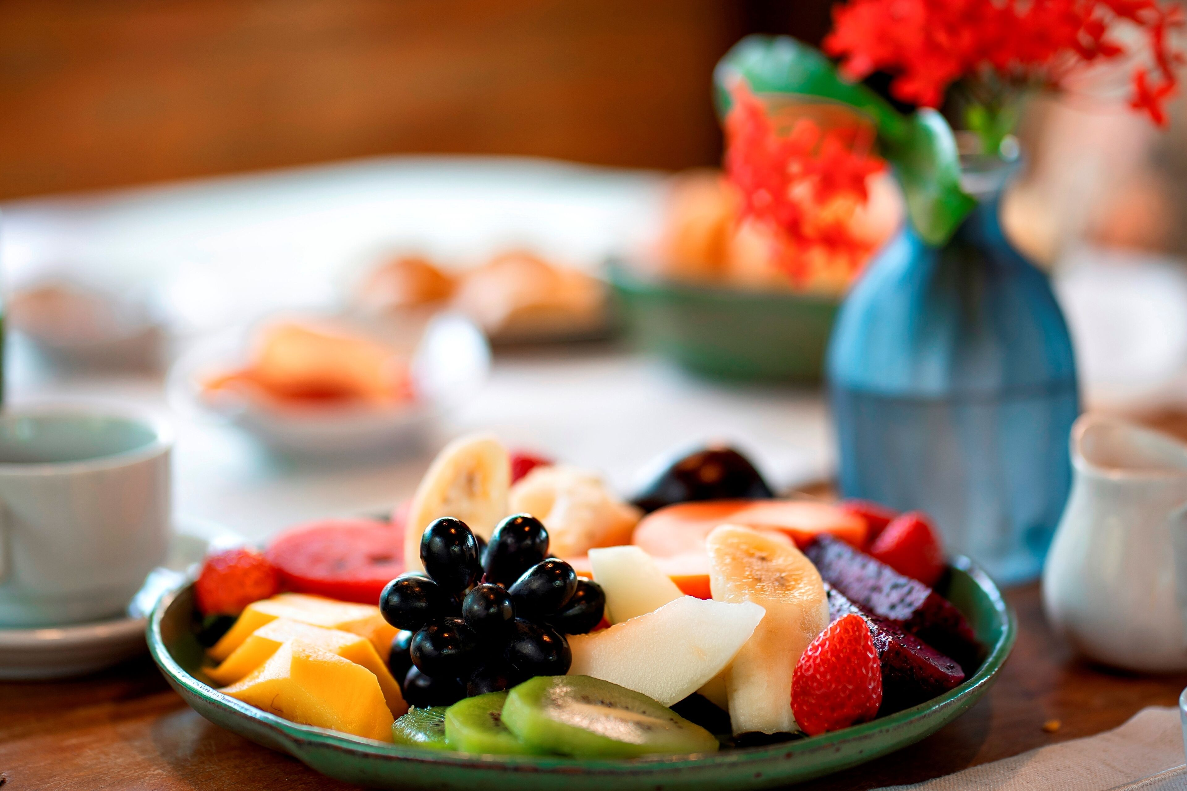 Una bandeja de frutas sobre una mesa, con un jarrón de flores al fondo.