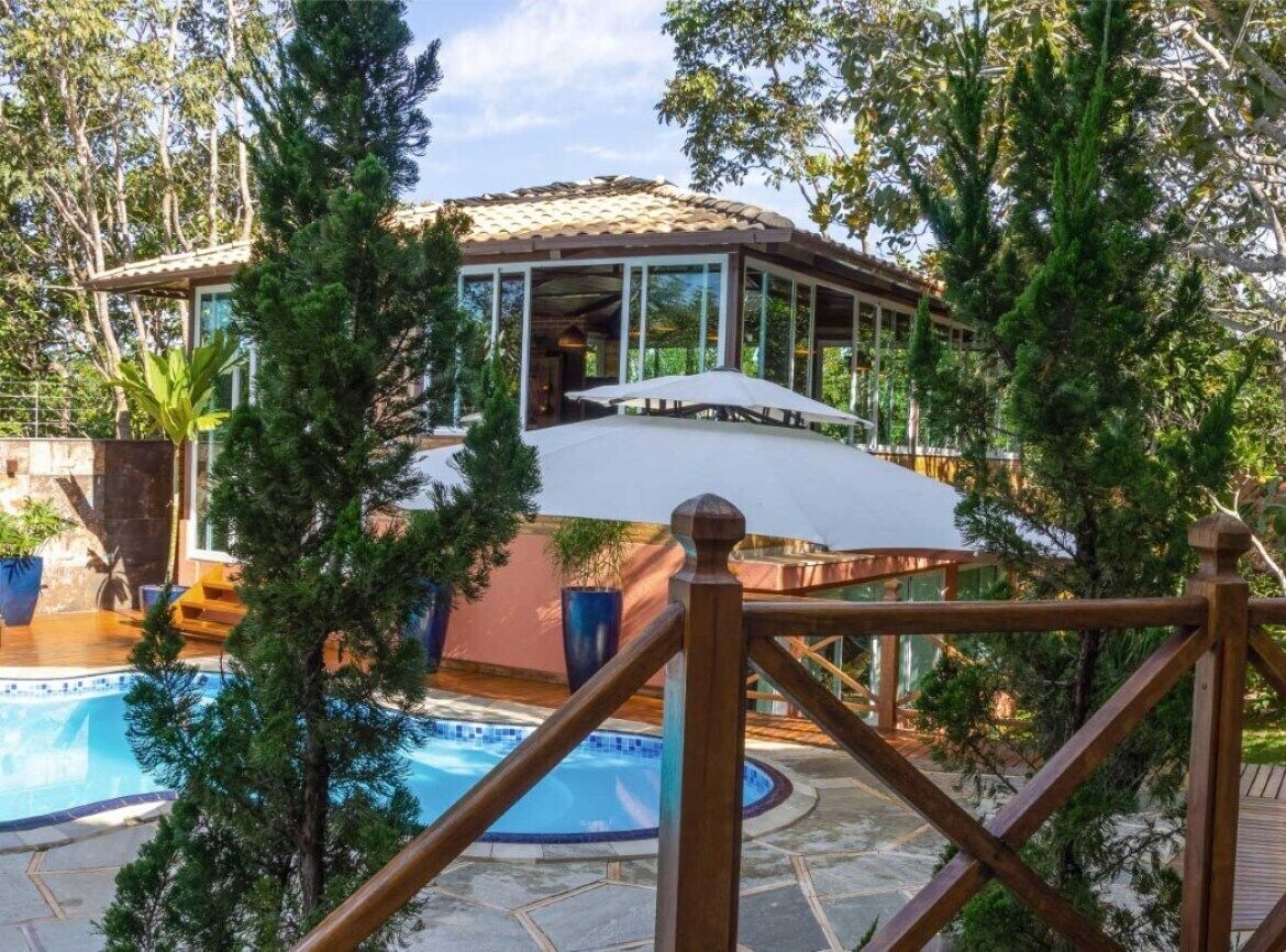 Zona de piscina con área de descanso cubierta, piscina y vegetación circundante.