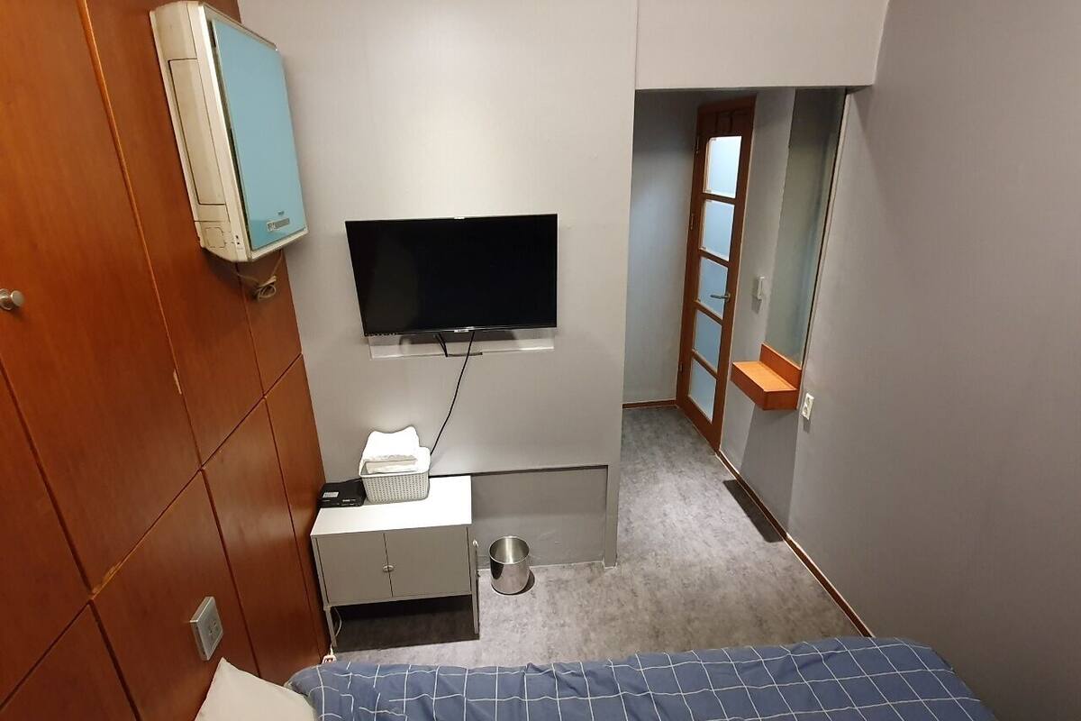 Habitación de hotel pequeña con cama, televisor montado en la pared, escritorio con lámpara y zapatero.