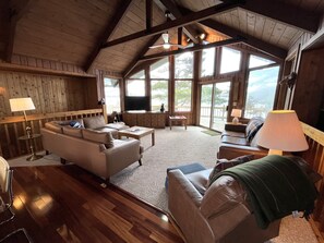 A spacious living room with a high ceiling, wooden walls, and large windows offering a view of the outdoors.