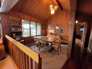 A wooden cabin interior with a dining table, chairs, a bookshelf, and a staircase.