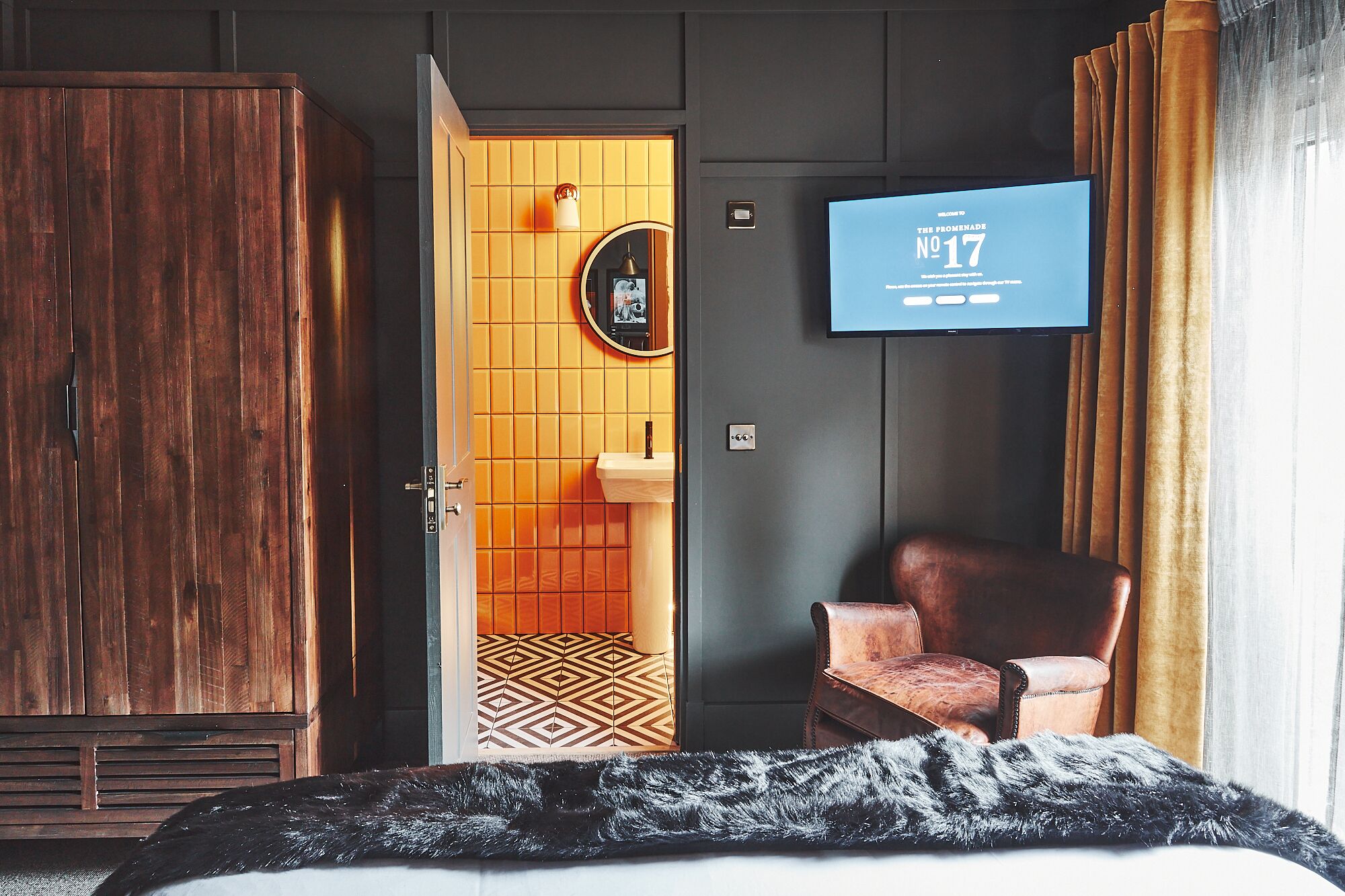 A hotel room with a bed, a television displaying 'Welcome to The Promenade No 17', a wooden wardrobe, a brown leather chair, and a patterned rug.