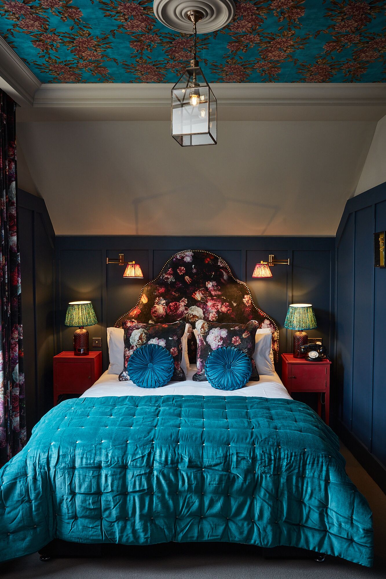 A bedroom with a teal bedspread, floral headboard, and two bedside lamps.