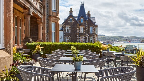 Een terras met rieten stoelen en tafels, een gebouw met stenen gevel in de achtergrond, en uitzicht op de stad.