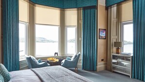 Een hotelkamer met een groot bed, twee fauteuils, een tafeltje en uitzicht op een waterpartij door grote ramen.