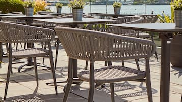 Terrasse extérieure avec des chaises et des tables en métal, des plantes en pot et une vue sur un lac et des collines en arrière-plan.