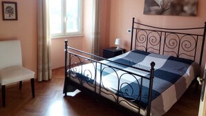 A bedroom with a metal bed frame, a bedside table with a lamp, a chair, a window with curtains, and a framed picture on the wall. - Large Vatican Apartment with 3 bedroom (Rome)
