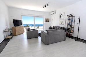 A modern living room with a grey sofa set, a television, a bookshelf, and a view of the sea through large windows.