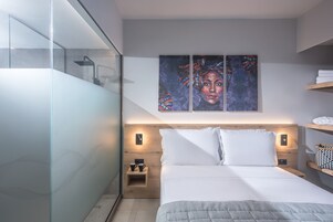 Habitación de hotel moderna con cabina de ducha de vidrio, cama con ropa blanca, cabecero de madera y una obra de arte colgada en la pared.