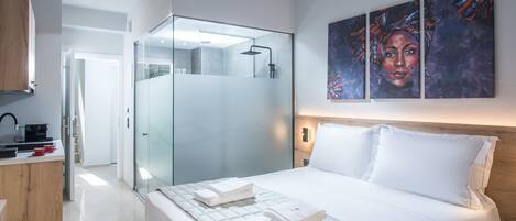 A modern hotel room with a large bed, a glass shower enclosure, a wooden desk, and a painting on the wall.